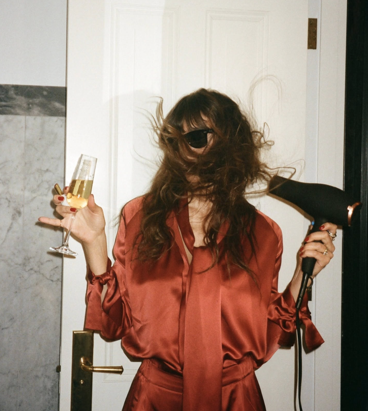 Woman in red silk holding champagne and a hair dryer, hair covering her face